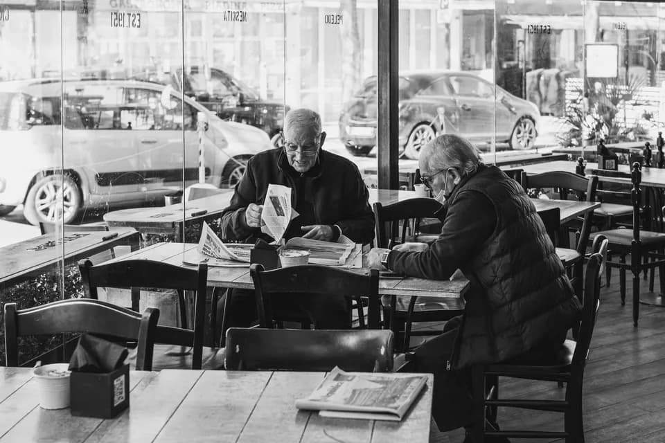 Two older men reading newspapers at a café table, in black and white.
