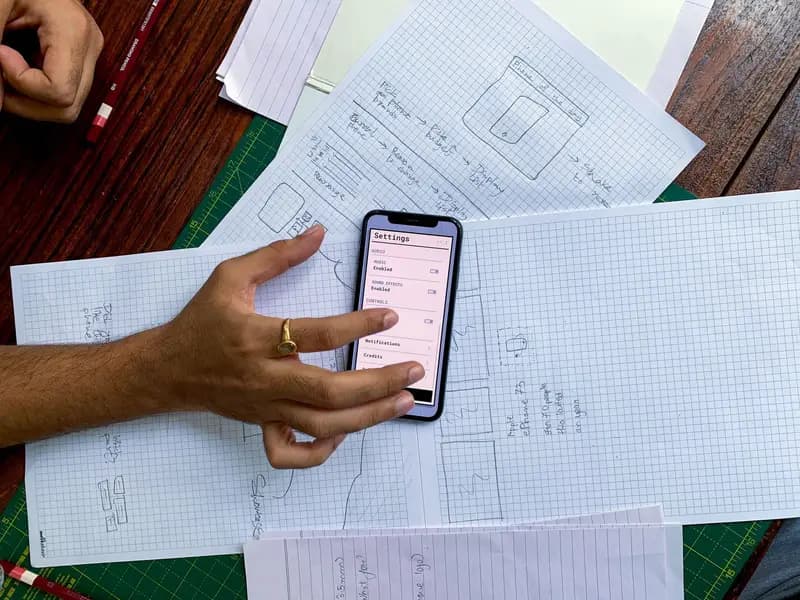 A hand holding a smartphone app screen over paper sketches of the same app's layout