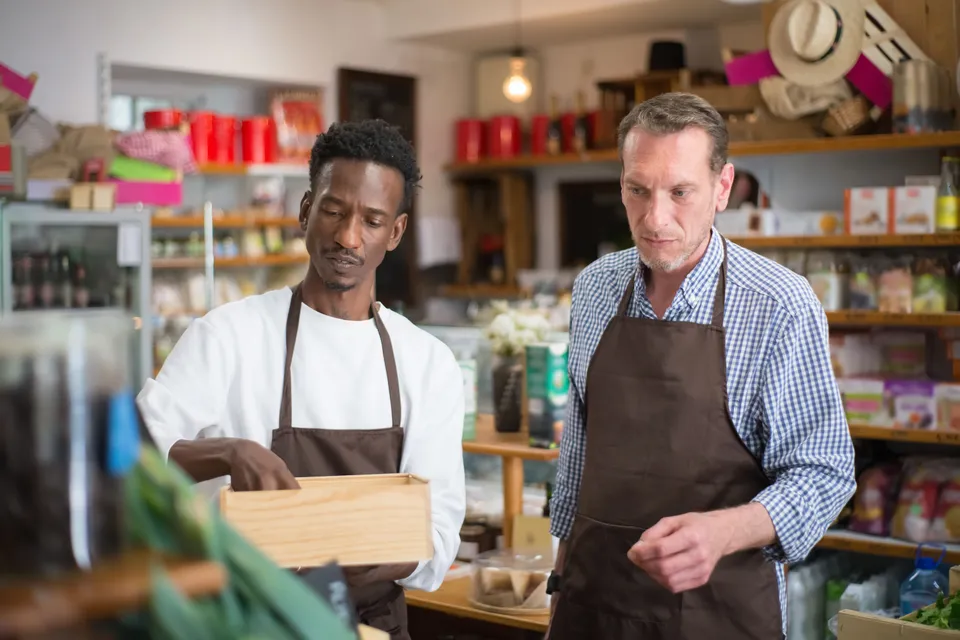 Two shopkeepers in aprons unpacking a crate of stock in a small grocery.