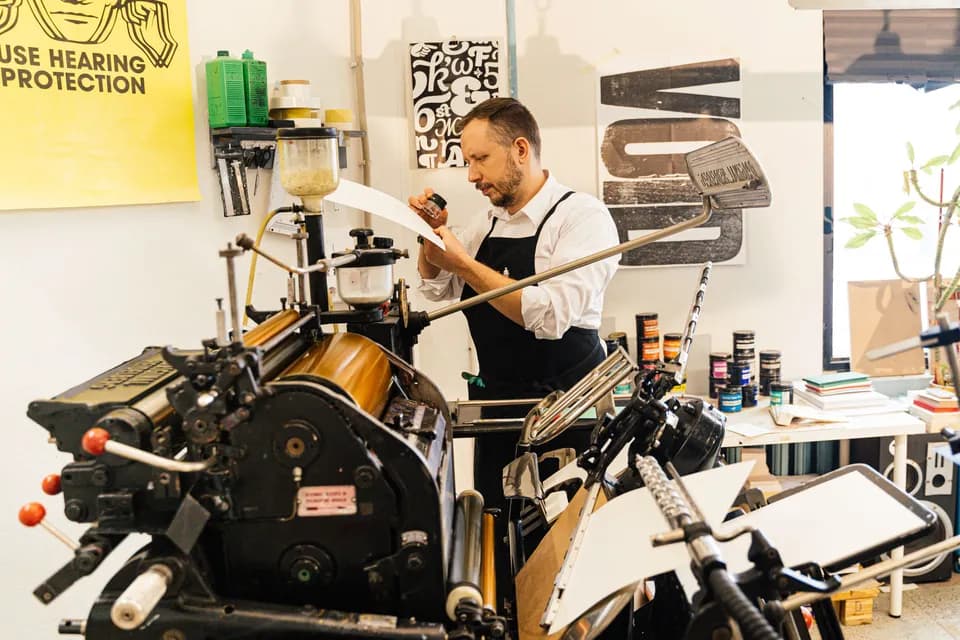 A printer in an apron checking a freshly printed sheet beside an old letterpress.