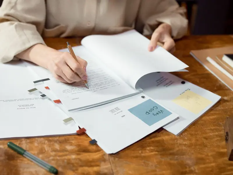 A hand marking up a printed manuscript with a pen, beside notes and colour swatches