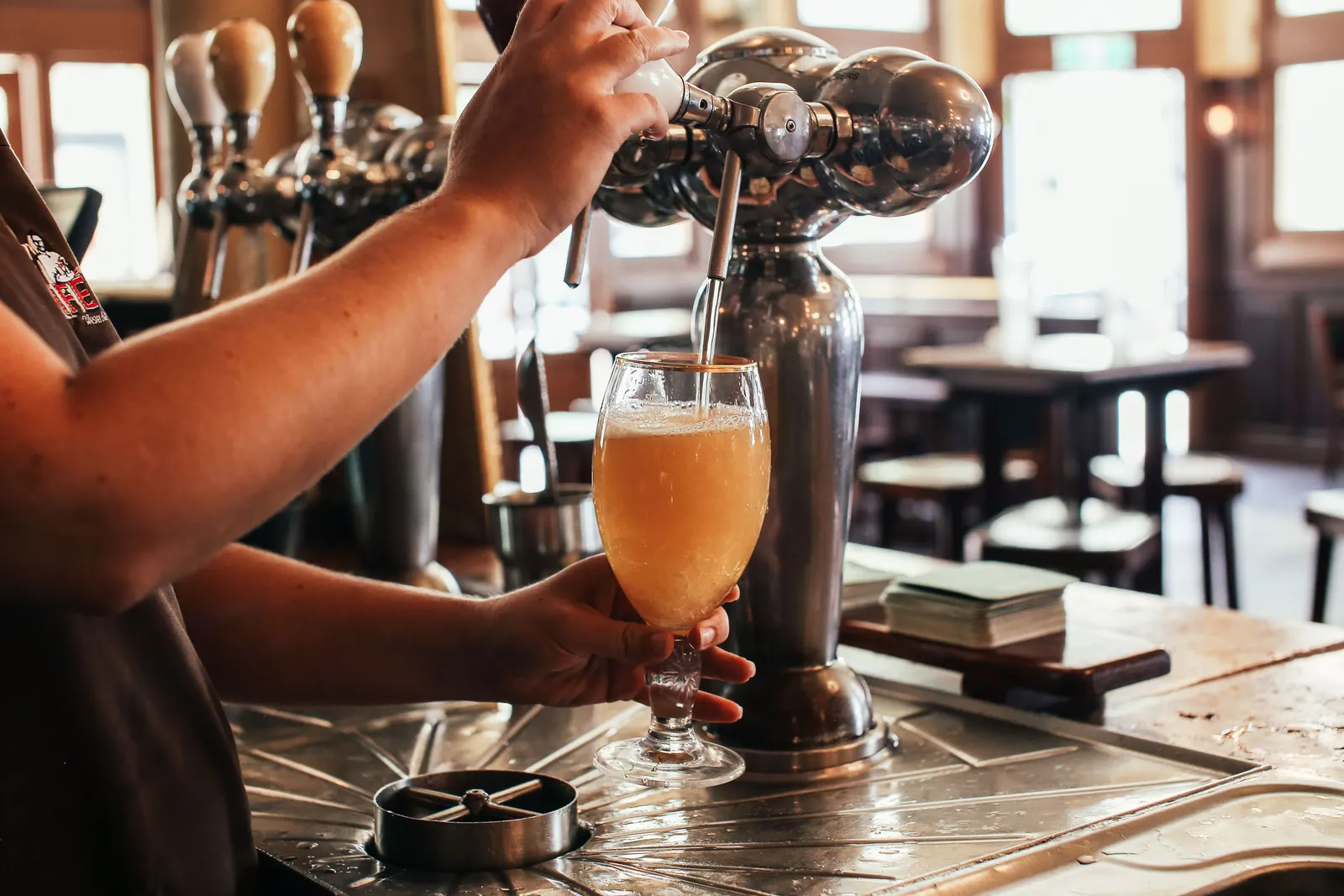 A bartender pulling a pint from a chrome tap on a pub bar, with empty tables and stools behind.