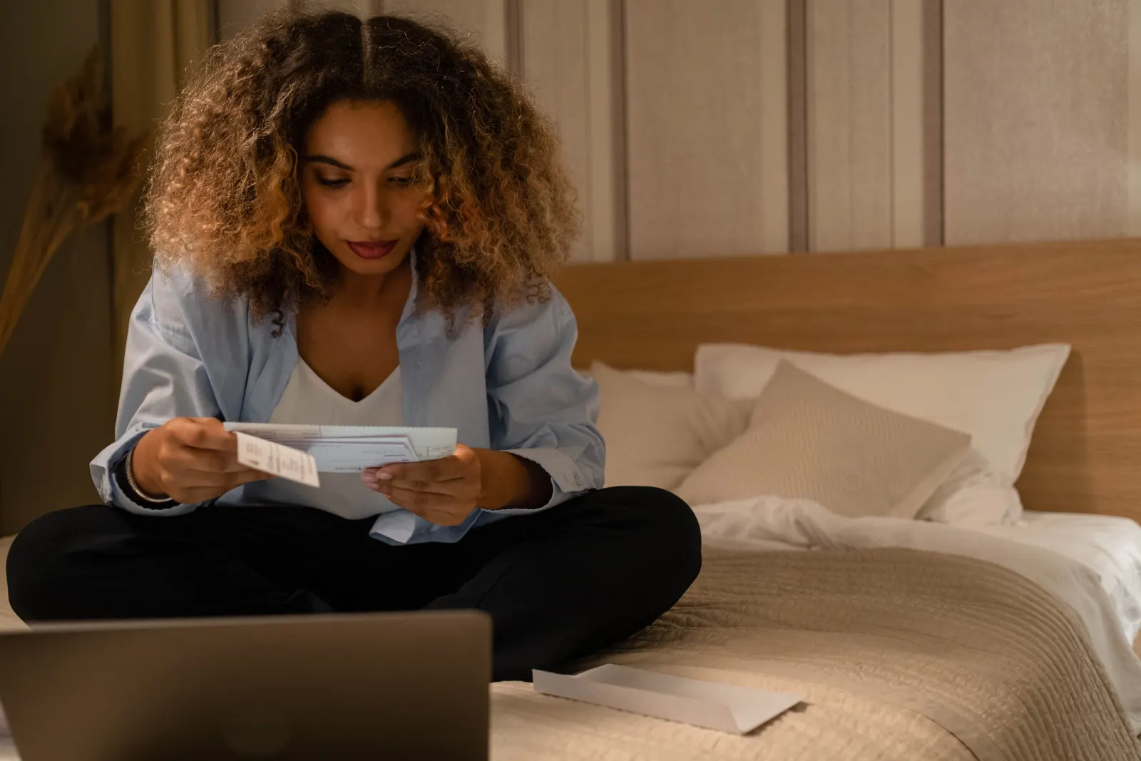 A woman sitting on her bed reading through a bill, with a laptop beside her.