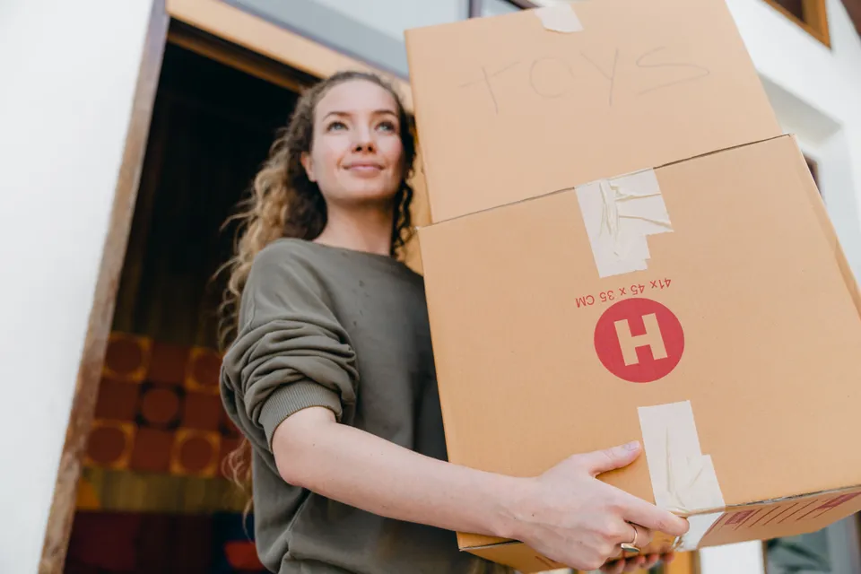A woman carrying two taped-up cardboard boxes out of a house on moving day.