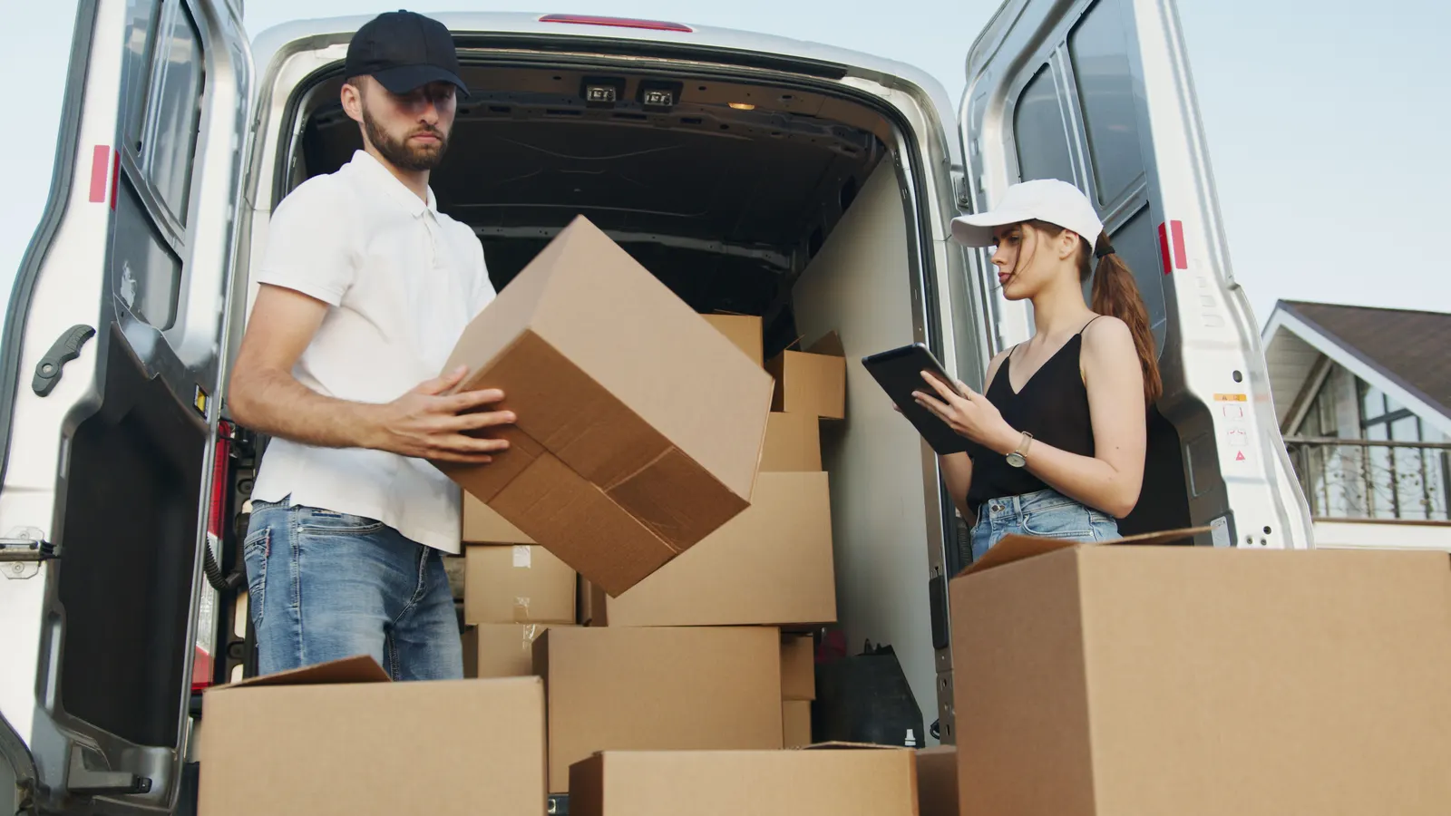 Two movers loading cardboard boxes into the back of a van while one checks a tablet.