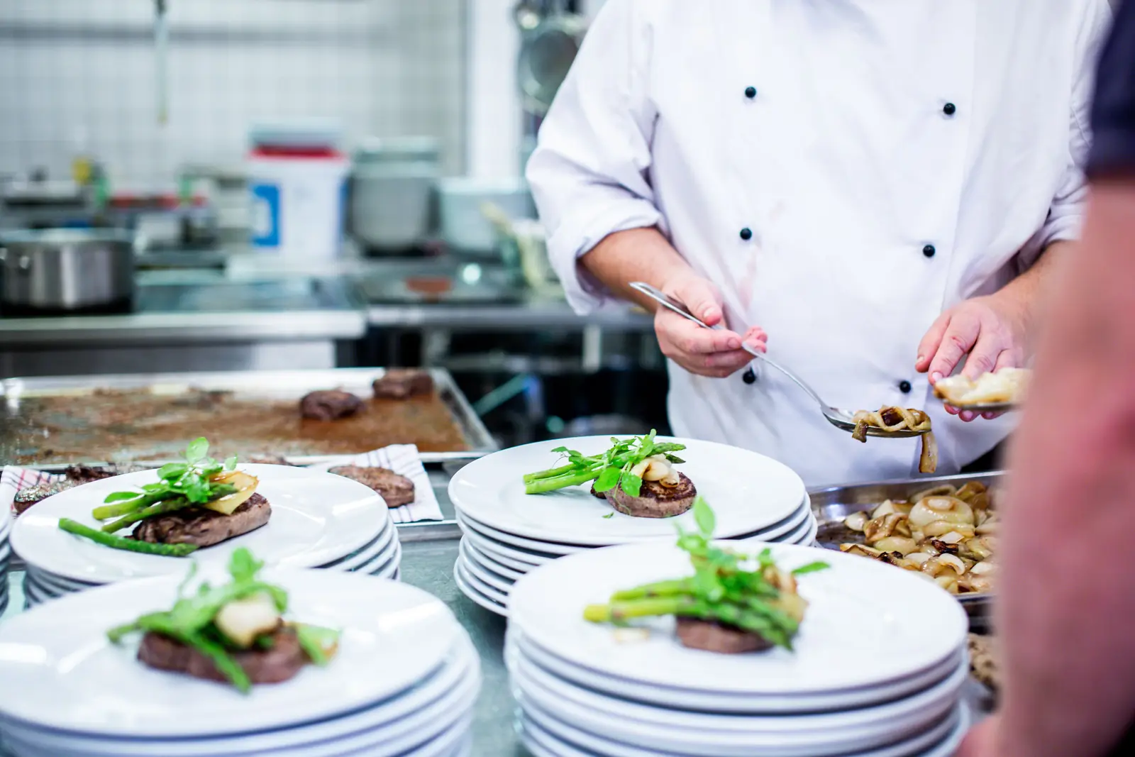 A chef spooning garnish onto a long row of identical plates at the pass during service.