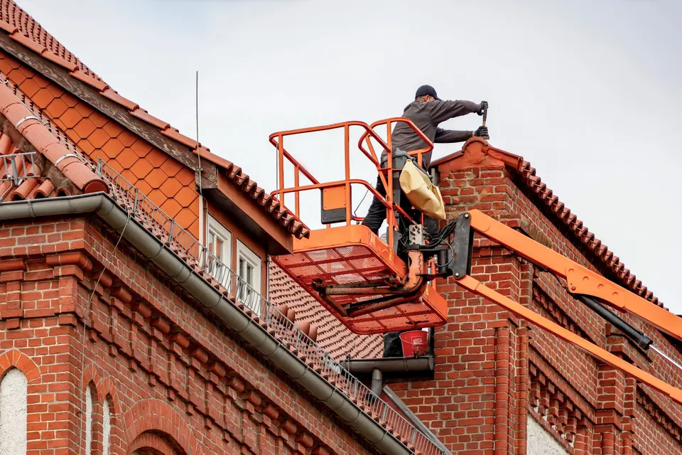 A roofer working from a cherry picker on the tiled roof of a red-brick building.
