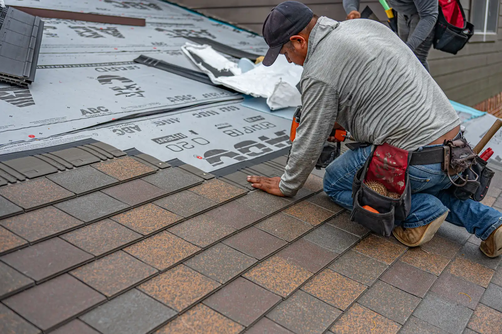 A roofer kneeling on a pitched roof, fixing new shingles in place.