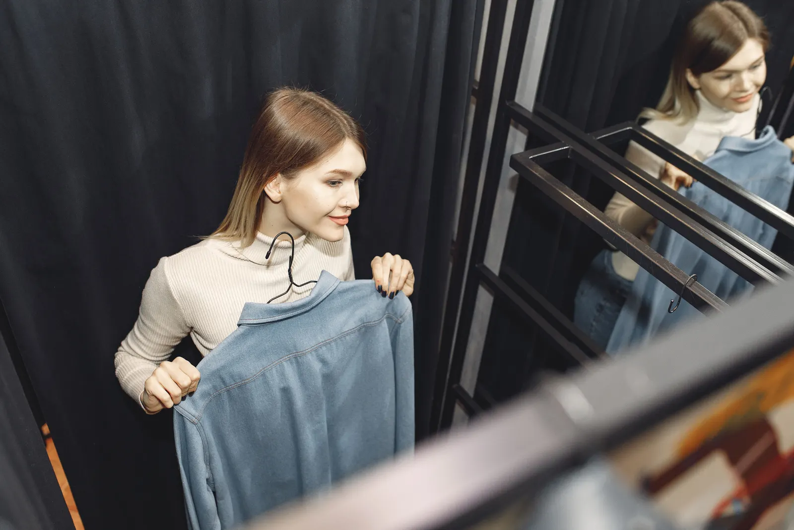 A woman holding up a jacket in a fitting room before deciding whether to buy it.