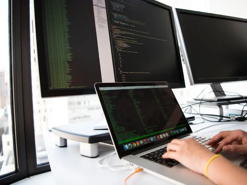 A developer typing on a laptop in front of two large monitors filled with code