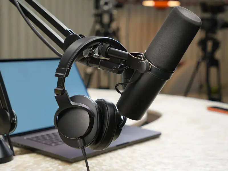 A podcast microphone on a boom arm with headphones hung over it, next to a laptop