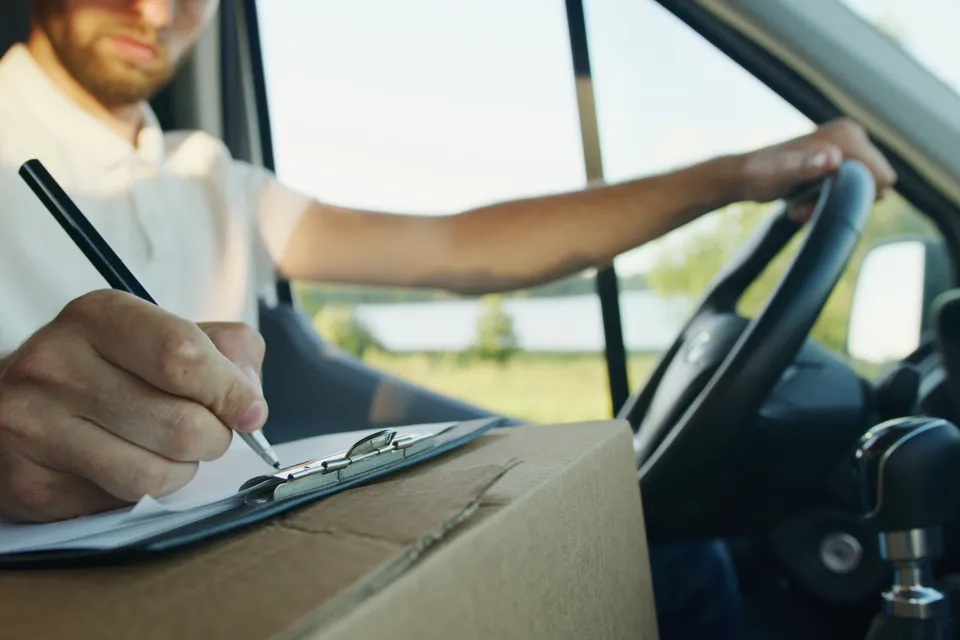 A driver sitting in his van, writing on a clipboard propped on a parcel.