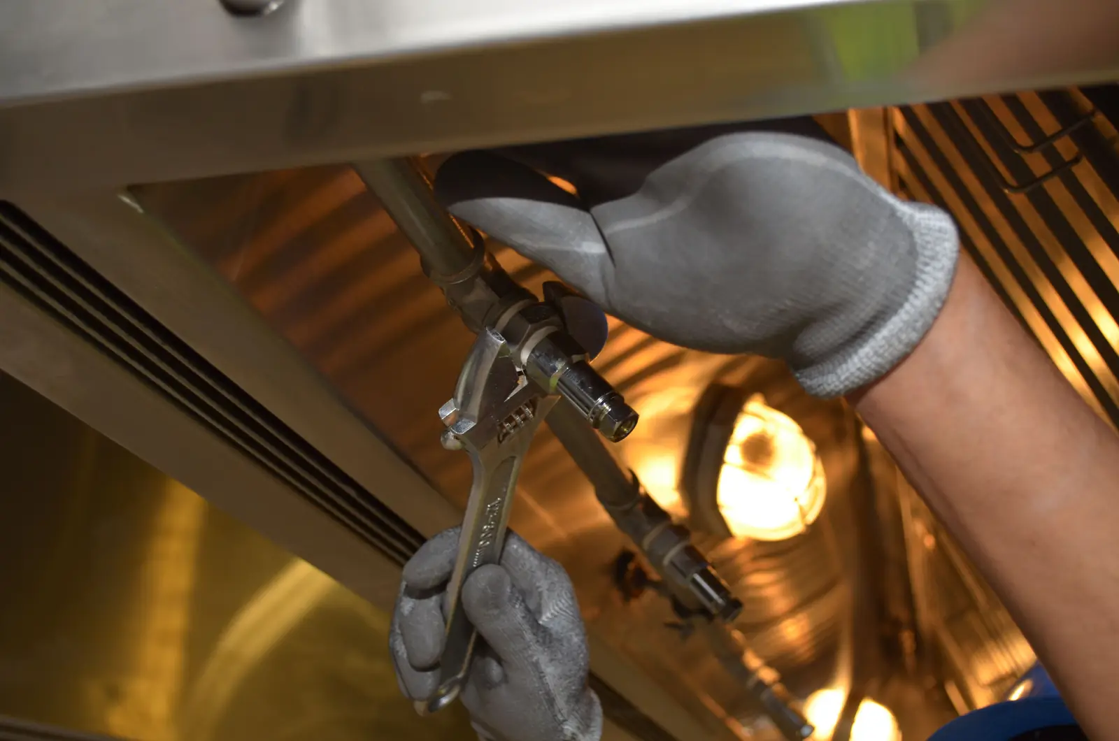 Gloved hands tightening a pipe fitting with an adjustable wrench, working overhead in a cramped space.