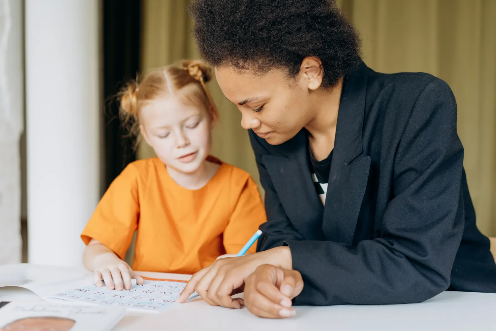 A tutor sitting beside a young pupil and helping with a writing exercise at a desk.