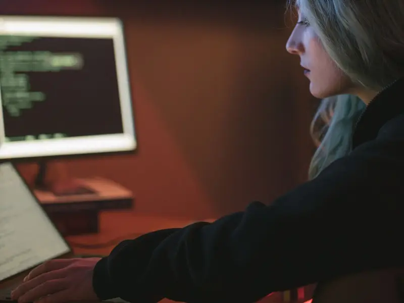 A person working late at a desk in a dark room, with code on the screens in front of her