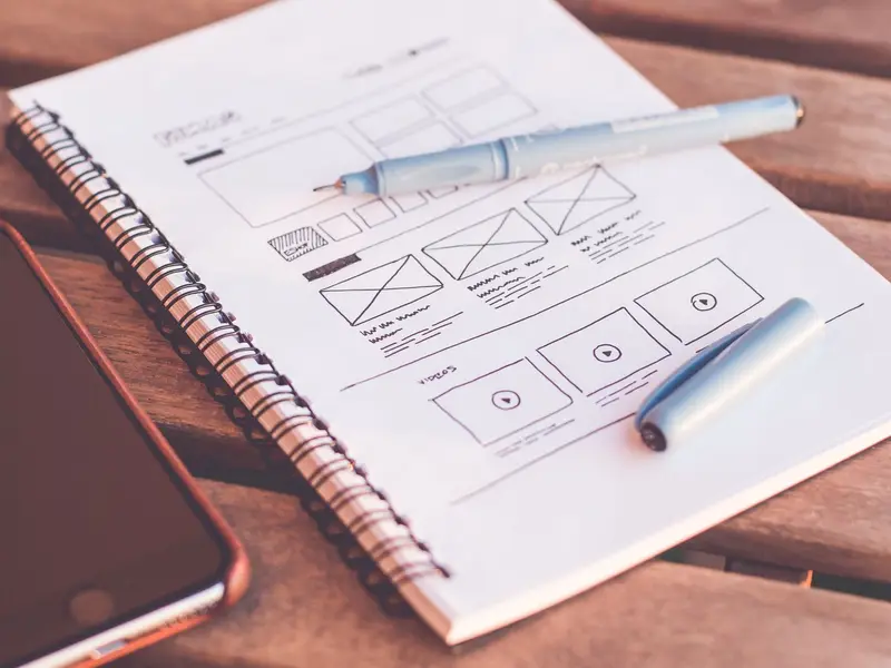 A notebook of page wireframe sketches with pens, beside a phone on a wooden desk