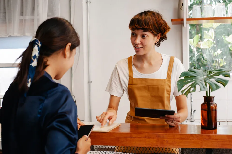 A café worker at the counter talking a colleague through something on a tablet.