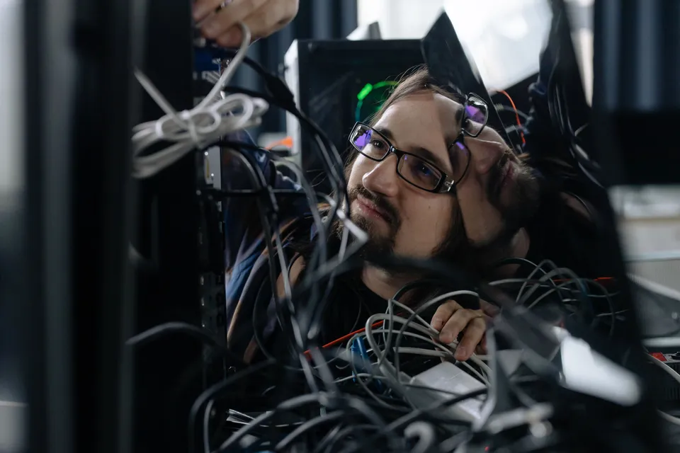 A technician peering out from behind a tangle of cables at the back of a workstation.