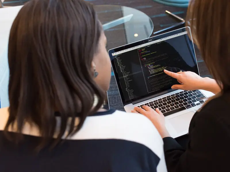 Two people looking at code on a laptop, one pointing at a line on the screen