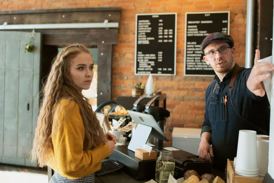 A barista at a café counter pointing a customer to the menu board behind him.