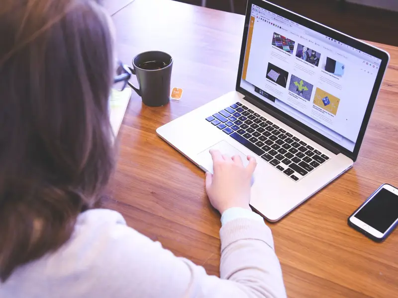 A person at a wooden desk with a website open on a laptop, a phone and a mug beside it