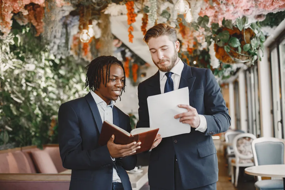Two men in suits going through a planning book in a venue decorated with flowers.