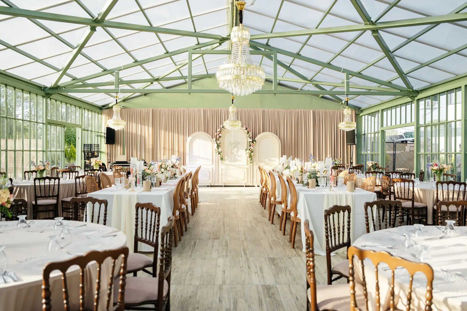 A glass-roofed venue laid for a reception, with round tables, chairs and chandeliers.