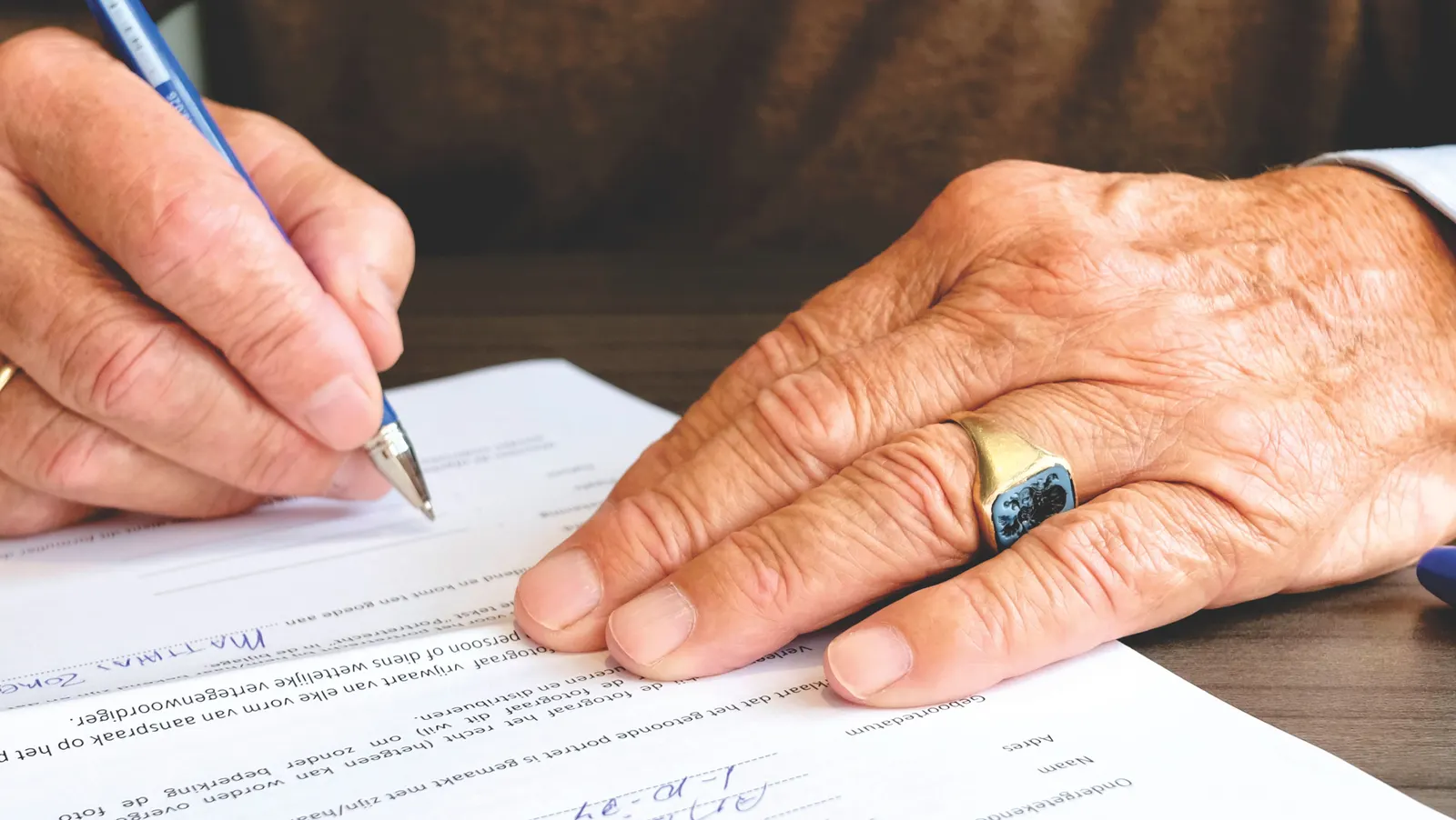 An older hand wearing a ring, signing a printed document with a pen.