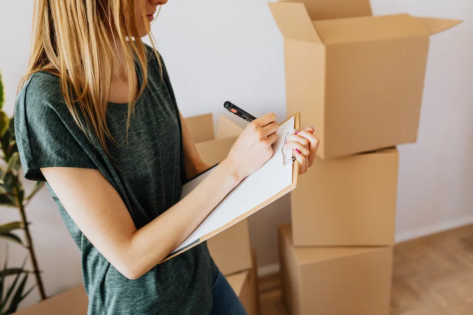 A woman ticking items off a list on a clipboard beside a stack of packed boxes.
